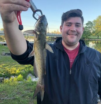 Picture of Tyler Butts, holding up a fish on a stringer in front of a small waterbody